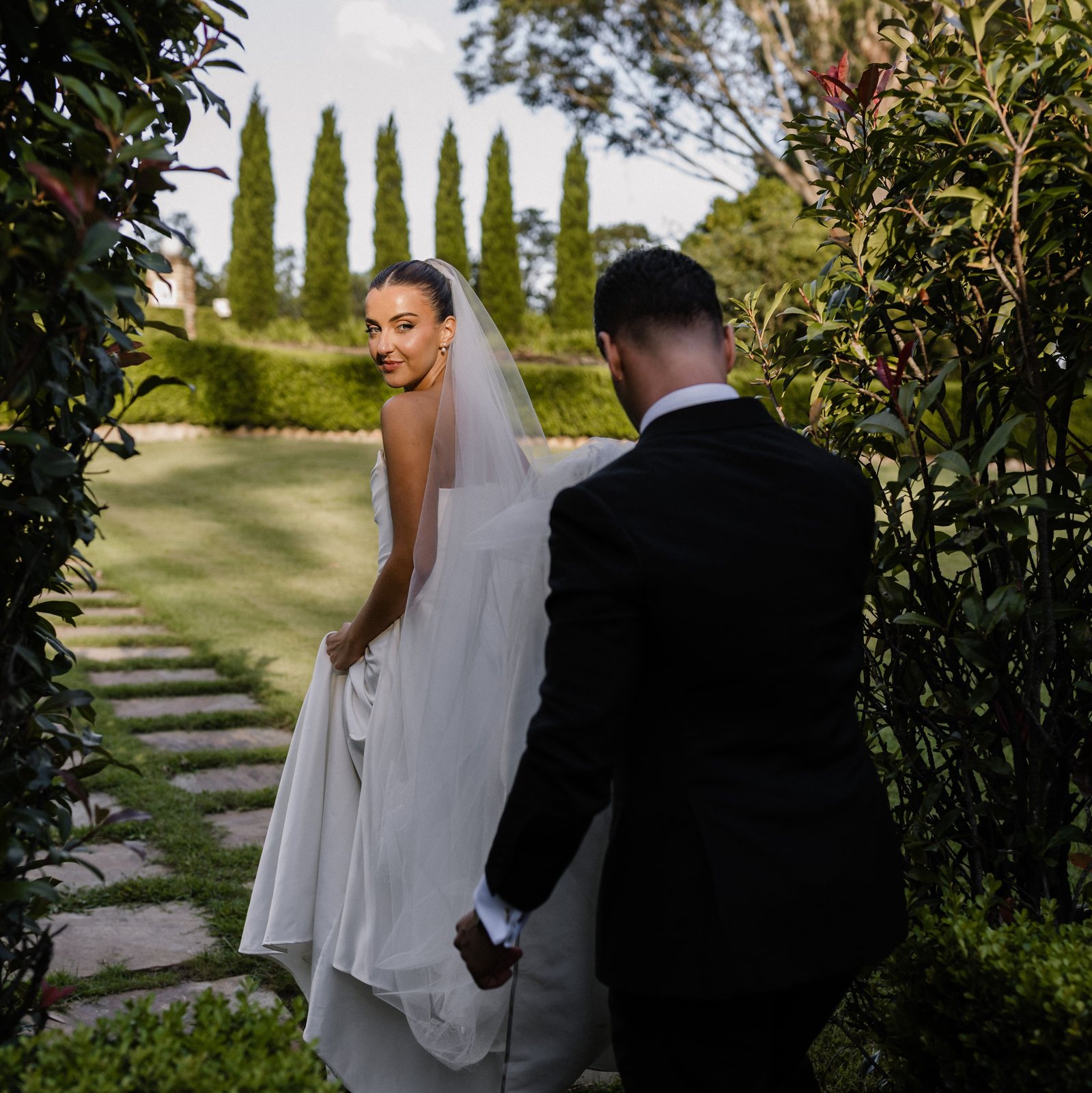 A bride looking back over her shoulder as the couple walk the stepping stones by the cypress hedge